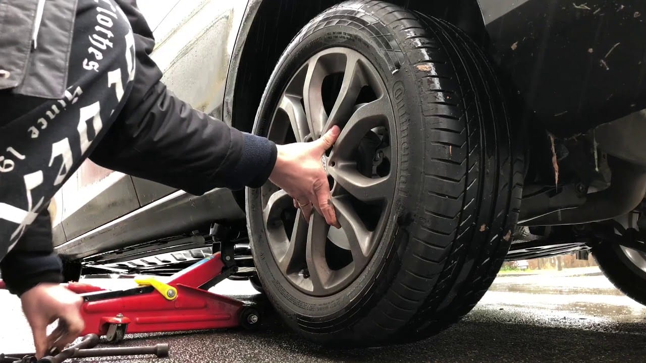 An auto mechanic uses a car jack to lift a vehicle while attempting to remove a wheel